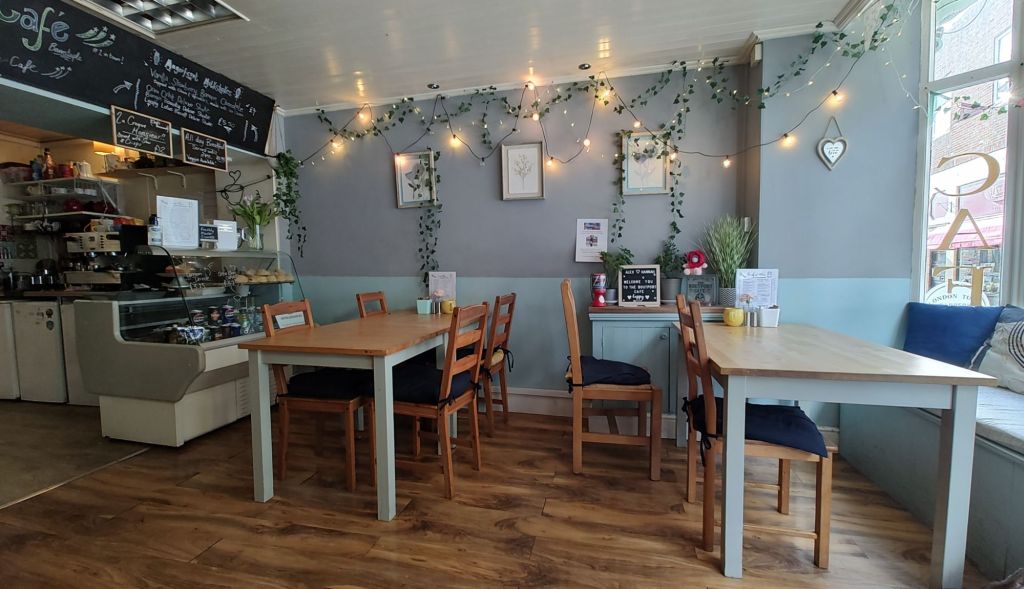 Interior of the Boutport Café showcasing simple wooden tables and chairs, with warm lighting and decorative plants. A chalkboard menu is displayed on the wall next to a counter with baked goods.