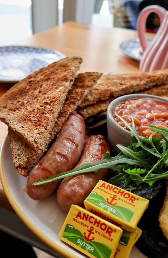 A plate featuring two grilled sausages, slices of whole grain toast, a small bowl of baked beans, greens, and butter portions on the side, set on a wooden table.