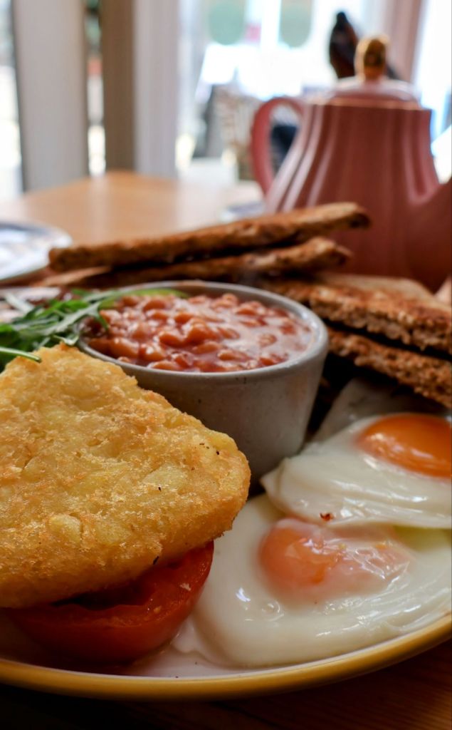 A hearty breakfast plate featuring two fried eggs, a hash brown, grilled tomato, baked beans in a small bowl, and slices of brown toast, set on a wooden table.