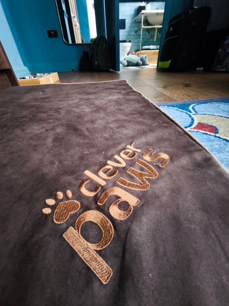 A close-up of a brown mat with the text 'clever paws' embroidered in gold, positioned in a stylish room with blue walls and glimpses of a bathroom in the background.