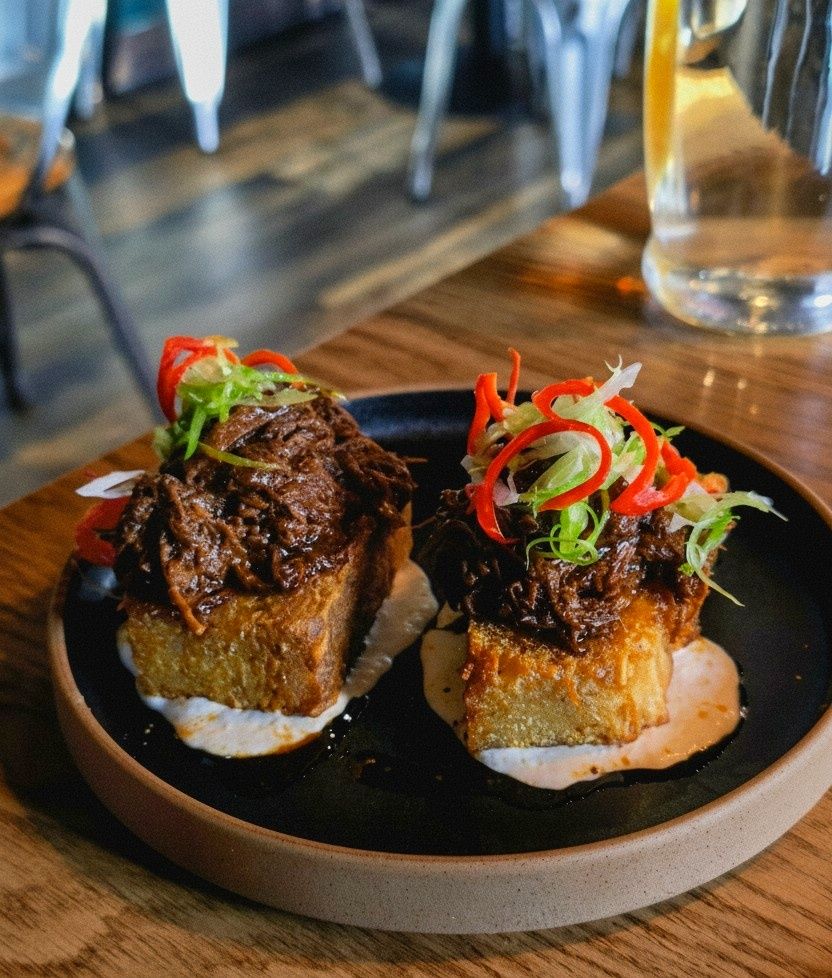 A close-up of two pieces of fried bread topped with pulled beef, garnished with sliced red chili, green onions, and a drizzle of sauce, served on a black plate.