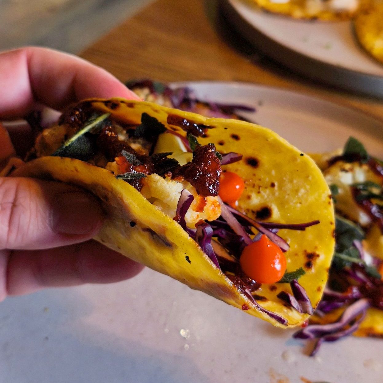 A hand holding a colorful taco filled with grilled vegetables, purple cabbage, and cherry tomatoes, with more tacos in the background.