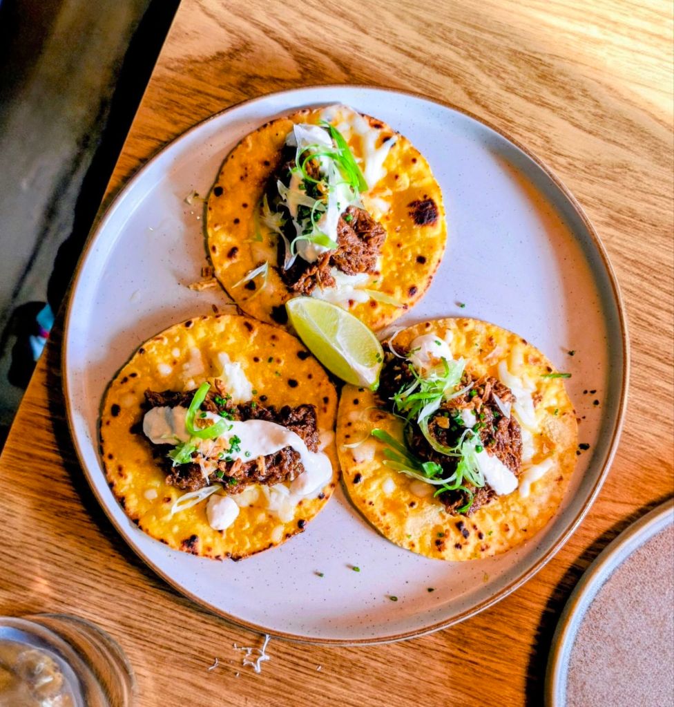 A plate of three tacos topped with beef, sour cream, and green onions, served with a lime wedge, on a wooden table.